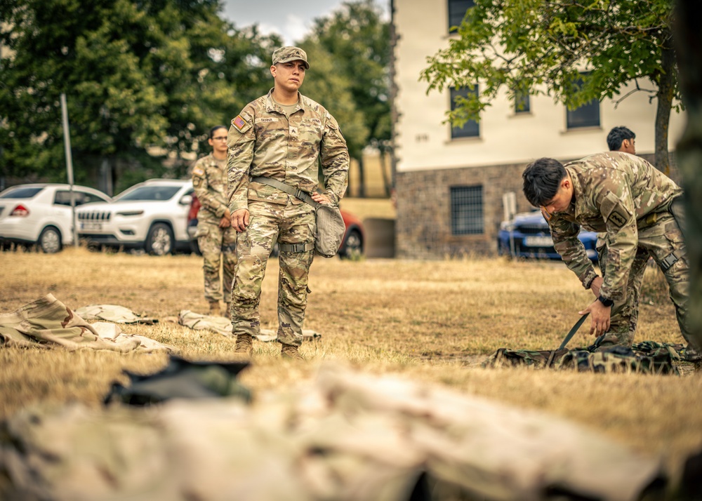 The image shows U.S. Army soldiers in MultiCam (OCP) uniforms conducting what appears to be a field equipment inspection or layout drill on a grassy area. At least four soldiers are visible, with one standing observantly while another bends down to handle gear. The setting appears to be a military installation or training area in Europe, suggested by the European-style architecture and vehicles in the background.