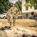 The image shows U.S. Army soldiers in MultiCam (OCP) uniforms conducting what appears to be a field equipment inspection or layout drill on a grassy area. At least four soldiers are visible, with one standing observantly while another bends down to handle gear. The setting appears to be a military installation or training area in Europe, suggested by the European-style architecture and vehicles in the background.