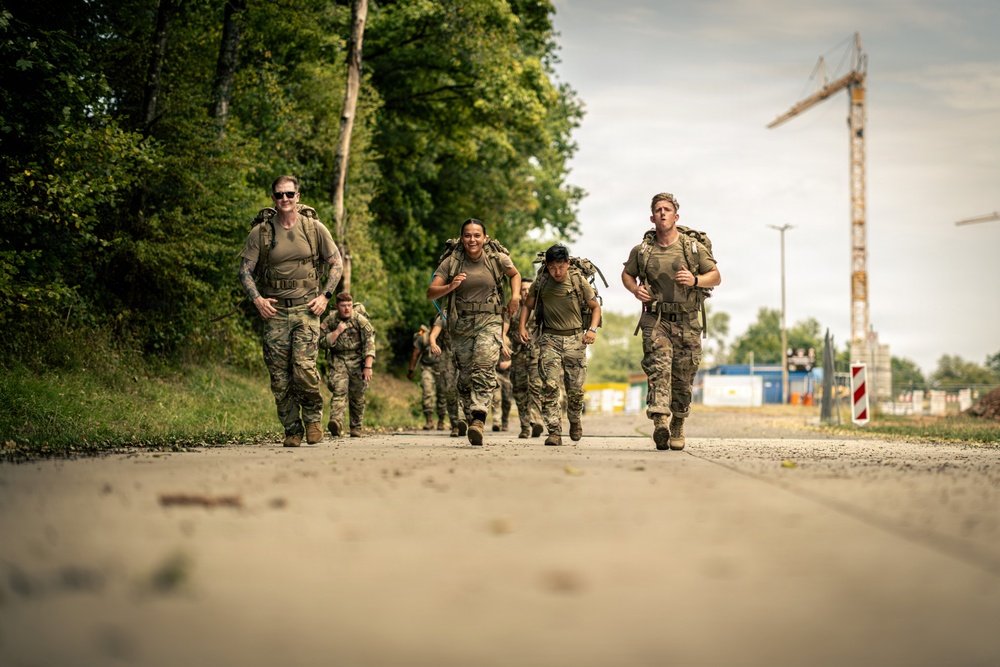A group of military personnel in camouflage uniforms are conducting a ruck march along a paved road, carrying tactical backpacks and gear. The group consists of both male and female soldiers moving at a brisk pace toward the camera. The setting appears to be a military installation or training area in a European location, evidenced by the construction crane and surrounding infrastructure in the background.