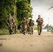 A group of military personnel in camouflage uniforms are conducting a ruck march along a paved road, carrying tactical backpacks and gear. The group consists of both male and female soldiers moving at a brisk pace toward the camera. The setting appears to be a military installation or training area in a European location, evidenced by the construction crane and surrounding infrastructure in the background.