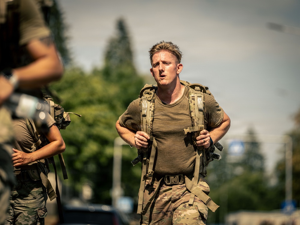A male soldier in military physical training attire is running or marching at pace while wearing a tactical rucksack/backpack. He appears physically exerted, with sweat visible on his olive-drab t-shirt and multicam trousers. At least one other similarly equipped individual is partially visible in the foreground left.