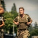 A male soldier in military physical training attire is running or marching at pace while wearing a tactical rucksack/backpack. He appears physically exerted, with sweat visible on his olive-drab t-shirt and multicam trousers. At least one other similarly equipped individual is partially visible in the foreground left.