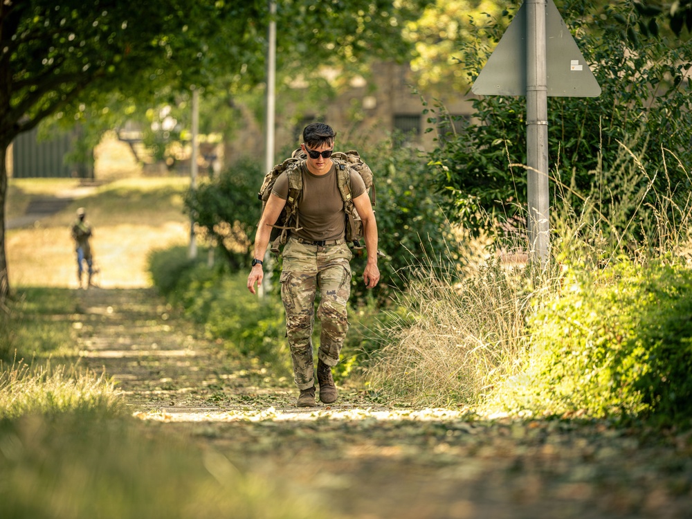 A soldier or military personnel in full camouflage uniform and tactical gear is walking along a tree-lined path, carrying a heavy rucksack. The image appears to capture a ruck march or physical training exercise in a semi-urban or base environment. A second similarly dressed individual is visible in the blurred background.