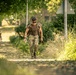 A soldier or military personnel in full camouflage uniform and tactical gear is walking along a tree-lined path, carrying a heavy rucksack. The image appears to capture a ruck march or physical training exercise in a semi-urban or base environment. A second similarly dressed individual is visible in the blurred background.