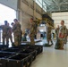 Military personnel in camouflage uniforms are gathered inside a large hangar or warehouse facility. Several soldiers appear to be receiving instructions or conducting an inspection, with one individual holding a red folder/clipboard. The space contains stacked black equipment cases, duffel bags, and various supplies, suggesting a deployment or logistics operation.