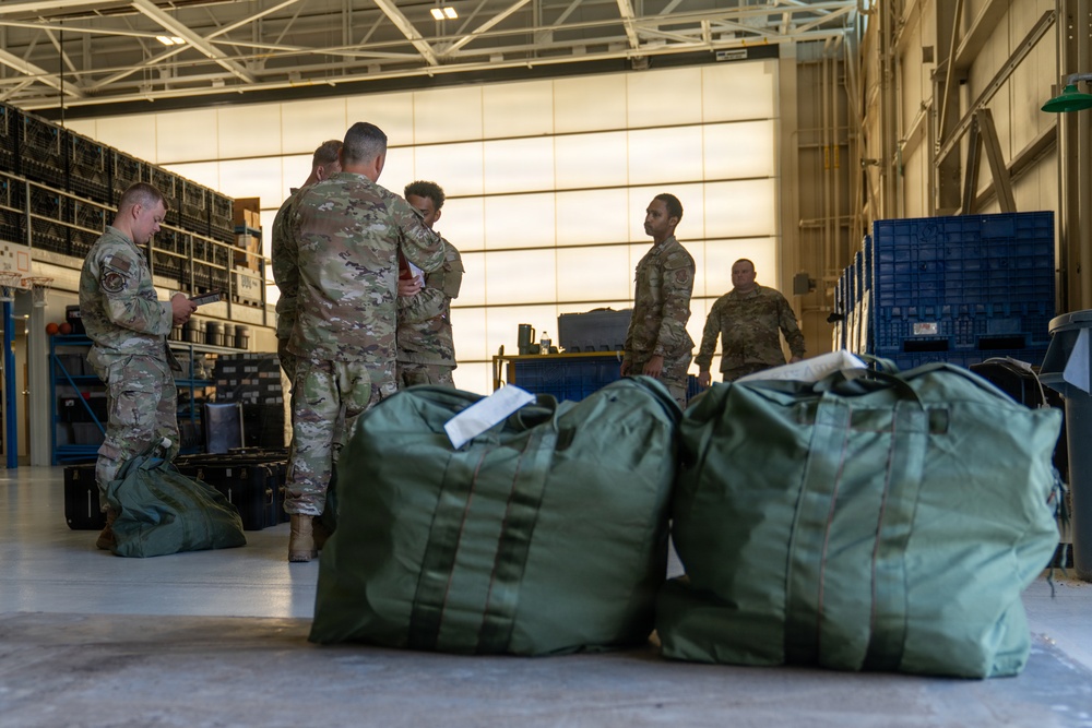 Military personnel in OCP (Operational Camouflage Pattern) uniforms are gathered inside what appears to be a large hangar or warehouse facility. Several large olive-drab military duffel bags are staged in the foreground, suggesting a deployment, equipment staging, or logistics operation. The personnel appear to be reviewing documents and coordinating tasks.
