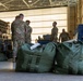 Military personnel in OCP (Operational Camouflage Pattern) uniforms are gathered inside what appears to be a large hangar or warehouse facility. Several large olive-drab military duffel bags are staged in the foreground, suggesting a deployment, equipment staging, or logistics operation. The personnel appear to be reviewing documents and coordinating tasks.