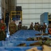 Military personnel in camouflage uniforms are working inside a large industrial hangar or warehouse facility. They appear to be conducting an inventory, inspection, or equipment staging operation, with numerous blue plastic storage crates arranged in rows. At least one individual is holding a red clipboard/folder, suggesting documentation or accountability procedures are underway.