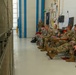 A group of military personnel in OCP (Operational Camouflage Pattern) uniforms are seated in folding chairs along a wall inside what appears to be a military hangar or maintenance facility. Several individuals are holding red folders or documents, suggesting a briefing, training session, or administrative review is taking place. One service member stands in the background near a blue-trimmed doorway, possibly in an observational or instructional role.