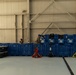 The image shows the interior of a large industrial hangar or warehouse facility with numerous blue plastic storage crates stacked in rows. Two individuals in military-style camouflage uniforms are visible among the crates, apparently conducting inventory or logistics work. Multiple pallet jacks in red, black, and yellow are positioned in front of the crate arrangement.