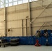 Two military personnel in camouflage uniforms are reviewing documents or a clipboard inside a large aircraft hangar. The hangar floor is lined with numerous blue plastic storage crates and pallets containing military gear, equipment cases, and supplies. The facility appears to be used for logistics staging or equipment inventory operations.