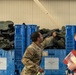 Two military personnel in MultiCam/OCP camouflage uniforms are conducting an inventory or inspection of chemical protective boots stored in large blue plastic crates. The crates are labeled 'BOOTS, CHEMICAL' with size designations (Size 13 and Size 14 visible). One soldier is physically handling the boots while the other holds a red binder and documents, suggesting an official inventory or logistics operation.