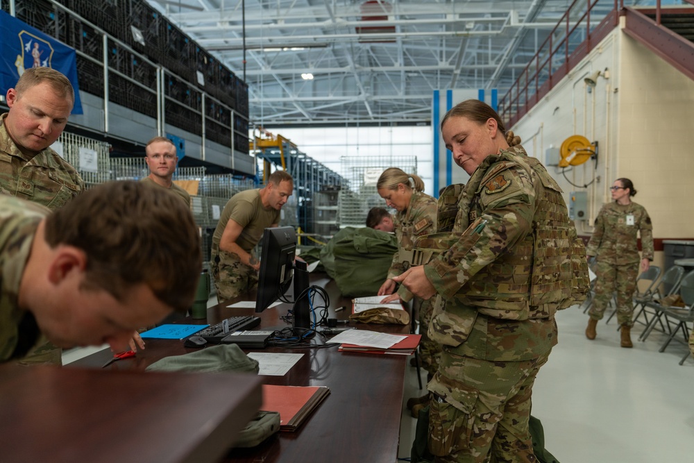 Military personnel in OCP (Operational Camouflage Pattern) uniforms are gathered in a large hangar or warehouse facility, processing paperwork and equipment at tables. The scene appears to be a deployment line, personnel accountability check, or equipment issue/turn-in event. Multiple service members are engaged in administrative and logistical tasks simultaneously.