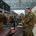 Military personnel in OCP (Operational Camouflage Pattern) uniforms are gathered in a large hangar or warehouse facility, processing paperwork and equipment at tables. The scene appears to be a deployment line, personnel accountability check, or equipment issue/turn-in event. Multiple service members are engaged in administrative and logistical tasks simultaneously.