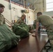 Military personnel in OCP (Operational Camouflage Pattern) uniforms are gathered around a long table in what appears to be a large indoor facility, likely a gymnasium or hangar. They appear to be conducting a gear inspection or equipment accountability check, with large olive drab duffel bags laid out on the table. The setting suggests a pre-deployment or mobilization processing event.
