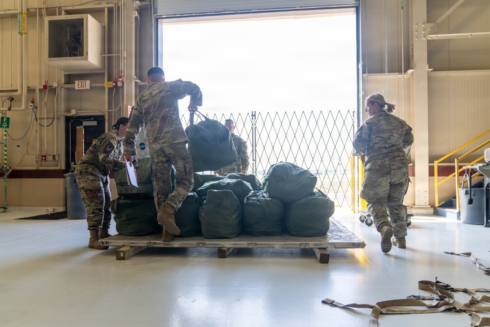 Military personnel in camouflage uniforms are working inside what appears to be a military hangar or loading facility, handling large olive-drab duffel bags stacked on a wooden pallet. The scene suggests a logistics or deployment operation, with soldiers loading, organizing, or inventorying gear near a large open bay door. The bright backlight from the open door creates a high-contrast environment.
