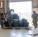 Military personnel in camouflage uniforms are working inside what appears to be a military hangar or loading facility, handling large olive-drab duffel bags stacked on a wooden pallet. The scene suggests a logistics or deployment operation, with soldiers loading, organizing, or inventorying gear near a large open bay door. The bright backlight from the open door creates a high-contrast environment.