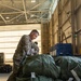 A U.S. Air Force service member in OCP (Operational Camouflage Pattern) uniform is inspecting or inventorying large olive drab military duffel bags stacked on a pallet inside what appears to be a military hangar or cargo facility. The service member is holding a document or label and appears to be checking tags on the bags. A second uniformed individual is visible in the background.