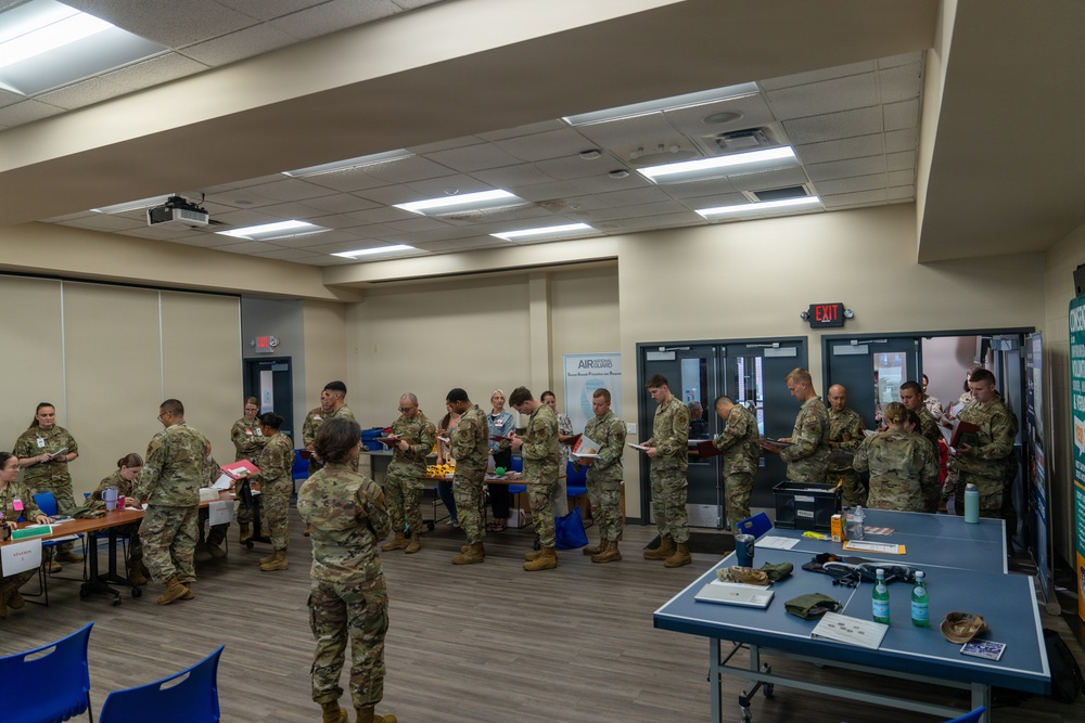 A group of military personnel in camouflage uniforms (OCP/MultiCam pattern) are gathered in what appears to be a briefing or processing room at a military facility. The personnel are standing in lines and reviewing documents or paperwork, suggesting an administrative processing event or pre-deployment briefing. A banner visible in the background references 'Air Guard' or similar Air National Guard branding.