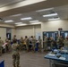 A group of military personnel in camouflage uniforms (OCP/MultiCam pattern) are gathered in what appears to be a briefing or processing room at a military facility. The personnel are standing in lines and reviewing documents or paperwork, suggesting an administrative processing event or pre-deployment briefing. A banner visible in the background references 'Air Guard' or similar Air National Guard branding.