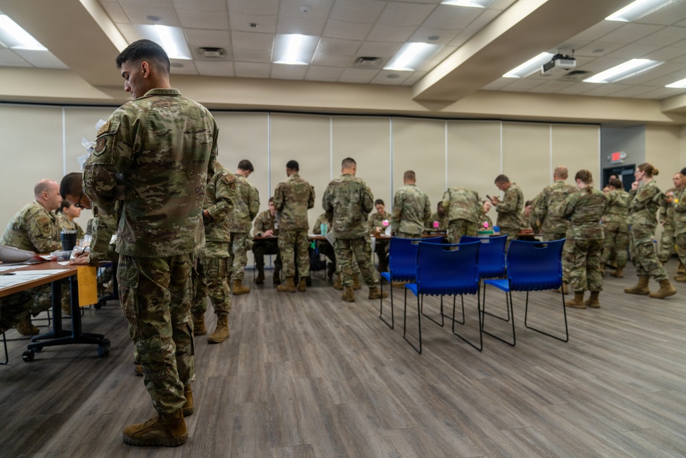 A large group of military personnel in OCP (Operational Camouflage Pattern) uniforms are gathered in what appears to be a conference or briefing room. The soldiers appear to be processing through administrative stations set up along tables. The setting suggests an in-processing, briefing, or administrative event at a military installation.
