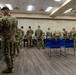 A large group of military personnel in OCP (Operational Camouflage Pattern) uniforms are gathered in what appears to be a conference or briefing room. The soldiers appear to be processing through administrative stations set up along tables. The setting suggests an in-processing, briefing, or administrative event at a military installation.