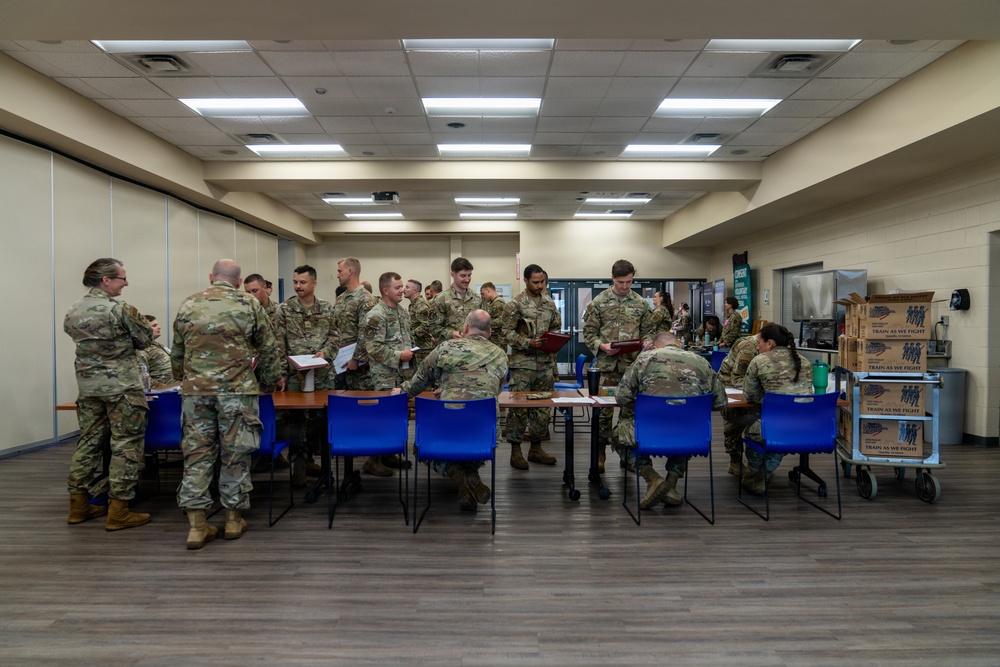 A group of military personnel in OCP (Operational Camouflage Pattern) uniforms are gathered in what appears to be a briefing or administrative processing area. Soldiers are standing and sitting around tables, reviewing documents and paperwork. The setting appears to be an indoor facility, possibly a community center or military building.