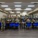 A group of military personnel in OCP (Operational Camouflage Pattern) uniforms are gathered in what appears to be a briefing or administrative processing area. Soldiers are standing and sitting around tables, reviewing documents and paperwork. The setting appears to be an indoor facility, possibly a community center or military building.