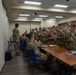 A group of military personnel in OCP (Operational Camouflage Pattern) uniforms are gathered in what appears to be a briefing or classroom facility. The soldiers appear to be engaged in some form of administrative processing, briefing, or training session, with some seated at tables and others standing in a queue-like formation. The setting is an indoor military facility with standard fluorescent lighting and blue chairs.
