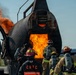 A group of firefighters in full protective gear and SCBA (Self-Contained Breathing Apparatus) units are conducting a live fire training exercise at what appears to be an aircraft rescue and firefighting (ARFF) training facility. The trainees are approaching a large metal aircraft fuselage simulator engulfed in active flames. One trainee is visibly wearing a jacket marked 'CRTC', indicating an organized training program.