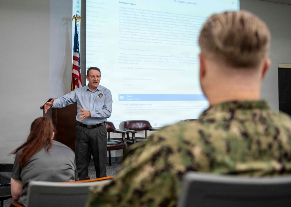 A civilian instructor or presenter is delivering a briefing or training session to a small audience that includes at least one uniformed U.S. Navy sailor in digital camouflage (NWU Type III) and a civilian attendee. The presentation screen behind the speaker displays a document titled 'HM - Hospital Corpsman' with a 'Rating Description' section visible. The setting appears to be a formal military or government conference/training room.