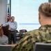 A civilian instructor or presenter is delivering a briefing or training session to a small audience that includes at least one uniformed U.S. Navy sailor in digital camouflage (NWU Type III) and a civilian attendee. The presentation screen behind the speaker displays a document titled 'HM - Hospital Corpsman' with a 'Rating Description' section visible. The setting appears to be a formal military or government conference/training room.