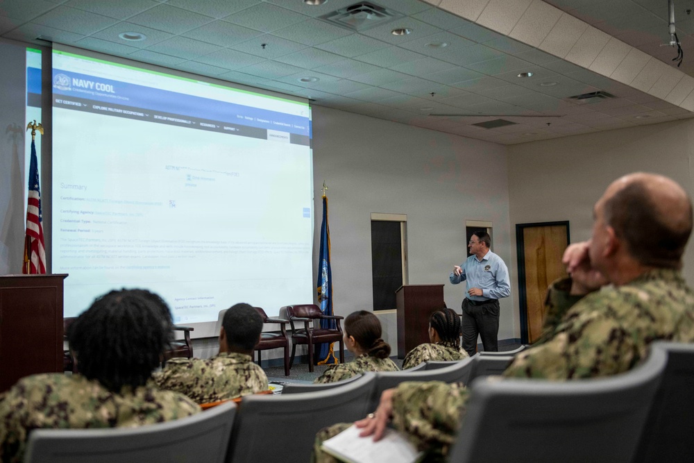 A civilian instructor is presenting a briefing on the 'Navy COOL' (Credentialing Opportunities On-Line) program to a group of U.S. Navy sailors in a conference or classroom setting. The projected screen displays the Navy COOL website, which helps service members obtain civilian certifications and credentials. The audience is seated in rows wearing Navy Working Uniform (NWU) camouflage pattern uniforms.