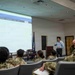 A civilian instructor is presenting a briefing on the 'Navy COOL' (Credentialing Opportunities On-Line) program to a group of U.S. Navy sailors in a conference or classroom setting. The projected screen displays the Navy COOL website, which helps service members obtain civilian certifications and credentials. The audience is seated in rows wearing Navy Working Uniform (NWU) camouflage pattern uniforms.
