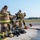Gulfport CRTC Live Fire Training Exercise A group of firefighters in tan/gold turnout gear with yellow reflective striping are preparing or debriefing at what appears to be an airfield or training facility. Several personnel are donning or doffing SCBA (Self-Contained Breathing Apparatus) equipment, with gear and fire hoses laid out on the ground. The setting suggests a training exercise or post-incident equipment check.