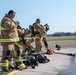 A group of firefighters in tan/gold turnout gear with yellow reflective striping are preparing or debriefing at what appears to be an airfield or training facility. Several personnel are donning or doffing SCBA (Self-Contained Breathing Apparatus) equipment, with gear and fire hoses laid out on the ground. The setting suggests a training exercise or post-incident equipment check.