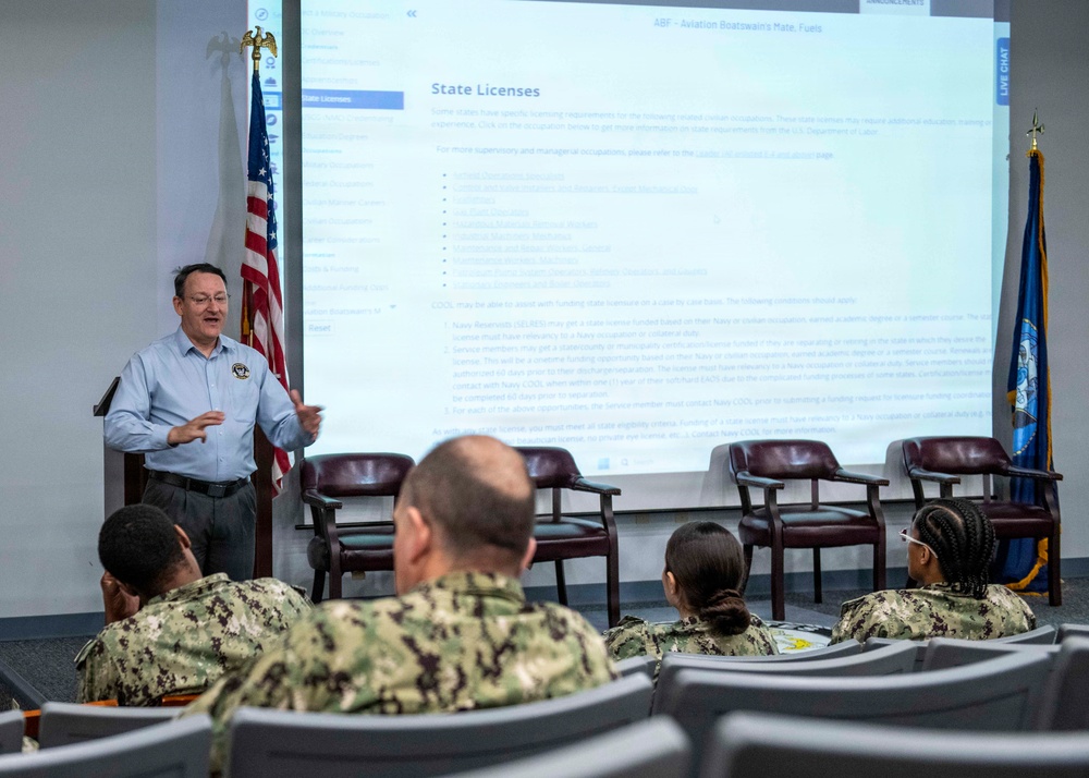 A civilian instructor or counselor is presenting a briefing on 'State Licenses' to a group of U.S. Navy sailors in a classroom or conference room setting. The projected screen displays information about state licensing requirements, referencing Navy COOL (Credentialing Opportunities On-Line) funding assistance. The audience consists of multiple sailors in Navy Working Uniform (NWU) camouflage pattern seated in rows.