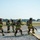 Gulfport CRTC Live Fire Training Exercise A group of firefighters in full turnout gear and SCBA (self-contained breathing apparatus) are conducting what appears to be a training exercise at an airfield or military base. They are working together to advance a fire hose across a gravel surface near a paved area with standing water. The setting, equipment, and coordinated team formation suggest this is an Aircraft Rescue and Firefighting (ARFF) or structural firefighting drill.