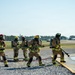 A group of firefighters in full turnout gear and SCBA (self-contained breathing apparatus) are conducting what appears to be a training exercise at an airfield or military base. They are working together to advance a fire hose across a gravel surface near a paved area with standing water. The setting, equipment, and coordinated team formation suggest this is an Aircraft Rescue and Firefighting (ARFF) or structural firefighting drill.
