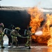 A team of firefighters in full protective gear and SCBA (self-contained breathing apparatus) are actively combating a large fire at what appears to be an aircraft fire training simulator. The firefighters are working in coordinated formation, directing a water/foam hose stream into intense orange flames engulfing the underside of a mock aircraft fuselage. The setting appears to be an airfield-based fire training facility, with standing water on the ground surface.