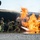 Gulfport CRTC Live Fire Training Exercise A team of firefighters in full protective gear are conducting what appears to be an aircraft rescue and firefighting (ARFF) training exercise. They are directing a hose line at large, active flames engulfing a mock aircraft fuselage structure on a paved training pad. The controlled environment, gravel surroundings, and purpose-built burn prop indicate this is a structured firefighting training scenario.