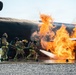 A team of firefighters in full protective gear are conducting what appears to be an aircraft rescue and firefighting (ARFF) training exercise. They are directing a hose line at large, active flames engulfing a mock aircraft fuselage structure on a paved training pad. The controlled environment, gravel surroundings, and purpose-built burn prop indicate this is a structured firefighting training scenario.