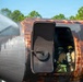 Firefighters in full protective gear and SCBA (self-contained breathing apparatus) are conducting a training exercise inside a mock aircraft fuselage. The heavily rusted and fire-scorched training prop simulates an aircraft cabin, with water being sprayed from the exterior on the left side. This appears to be an aircraft rescue and firefighting (ARFF) training drill at an outdoor training facility.