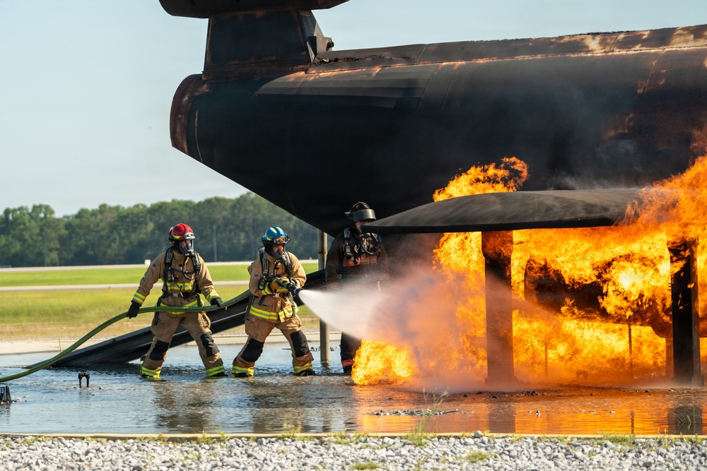 Two firefighters in full protective gear are actively combating a large fire engulfing what appears to be a mock aircraft fuselage structure, likely during a training exercise at an airfield. One firefighter wearing a red helmet and another wearing a blue helmet are directing a water hose at the base of intense orange flames. The setting includes a wet tarmac surface, gravel ground, and a tree line visible in the background, consistent with an airport fire training facility.