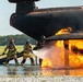 Two firefighters in full protective gear are actively combating a large fire engulfing what appears to be a mock aircraft fuselage structure, likely during a training exercise at an airfield. One firefighter wearing a red helmet and another wearing a blue helmet are directing a water hose at the base of intense orange flames. The setting includes a wet tarmac surface, gravel ground, and a tree line visible in the background, consistent with an airport fire training facility.