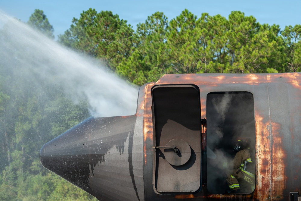 A firefighter in full protective gear is visible inside or near the doorway of a heavily rusted, burned aircraft fuselage during what appears to be a firefighting training exercise. A powerful water stream is being directed at the nose section of the aircraft from the left side. Smoke is visible emanating from the aircraft's windows and interior.