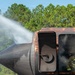 A firefighter in full protective gear is visible inside or near the doorway of a heavily rusted, burned aircraft fuselage during what appears to be a firefighting training exercise. A powerful water stream is being directed at the nose section of the aircraft from the left side. Smoke is visible emanating from the aircraft's windows and interior.