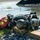 Gulfport CRTC Live Fire Training Exercise Firefighting equipment is laid out on a concrete curb edge near a body of water. The gear includes a Self-Contained Breathing Apparatus (SCBA) with carbon fiber air cylinders, a red firefighter helmet with high-visibility yellow-green markings, and black tactical gloves. The equipment appears to have been recently used, possibly during a water rescue or firefighting training exercise.