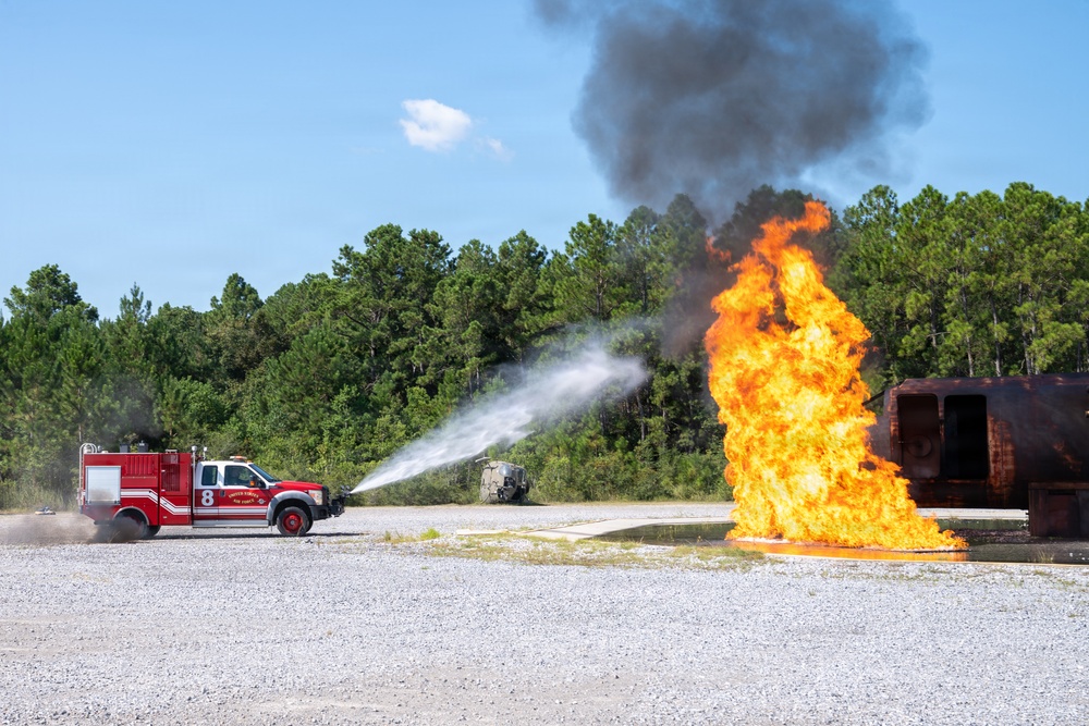 A red fire truck numbered '8' is actively discharging a high-pressure water stream toward a large open flame at what appears to be a firefighting training facility. The fire is burning intensely near a rusted, decommissioned aircraft or vehicle fuselage used as a training prop. Black and gray smoke rises into a clear blue sky above a gravel-covered open area surrounded by pine trees.