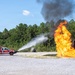 A red fire truck numbered '8' is actively discharging a high-pressure water stream toward a large open flame at what appears to be a firefighting training facility. The fire is burning intensely near a rusted, decommissioned aircraft or vehicle fuselage used as a training prop. Black and gray smoke rises into a clear blue sky above a gravel-covered open area surrounded by pine trees.